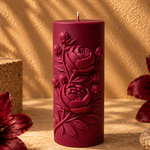 A dark burgundy cylindrical pillar candle with embossed floral relief, standing on a textured beige surface with warm sunlight and leaf shadows.